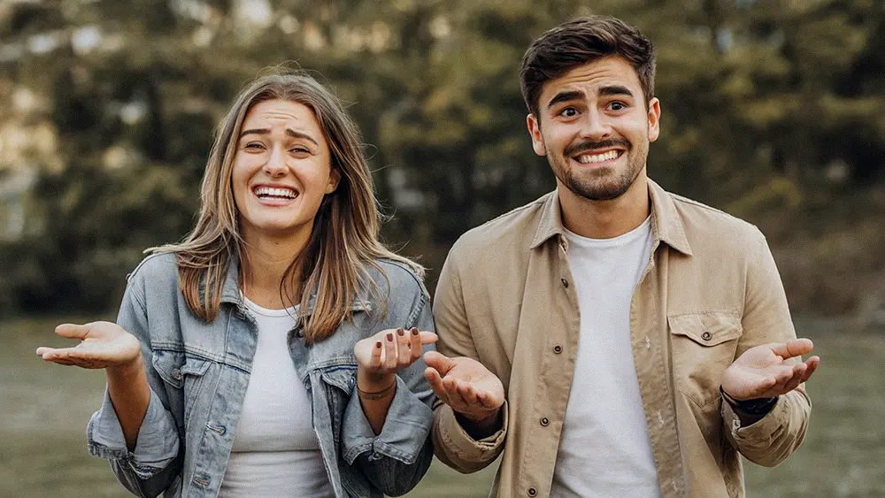 A young couple standing outdoors shrugging and smiling awkwardly, illustrating the mutual uncertainty of modern dating.